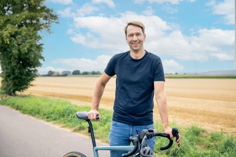Das Porträtfoto zeigt Jan Dingeldey in einer authentischen und aktiven Szenerie unter freiem Himmel. In einer entspannten Pose steht er auf einem asphaltierten Radweg, die Hände locker am Sattel und am Lenker eines modernen Gravelbikes positioniert. Jan Dingeldey trägt ein schlichtes, dunkelblaues T-Shirt und eine klassische Bluejeans, was den nahbaren und bodenständigen Charakter des Bildes unterstreicht. Das Fahrrad besticht durch ein minimalistisches Design in einem hellen Blauton und ist mit sportlichen Komponenten sowie profilierten Reifen für unbefestigte Wege ausgestattet. Im Hintergrund erstreckt sich eine weite, goldgelbe Agrarlandschaft mit einem abgeernteten Getreidefeld, das typisch für die Region ist. Ein großer, grüner Laubbaum am linken Bildrand und der weite, hellblaue Himmel mit lockerer Bewölkung verleihen der Aufnahme eine freundliche und sommerliche Atmosphäre.